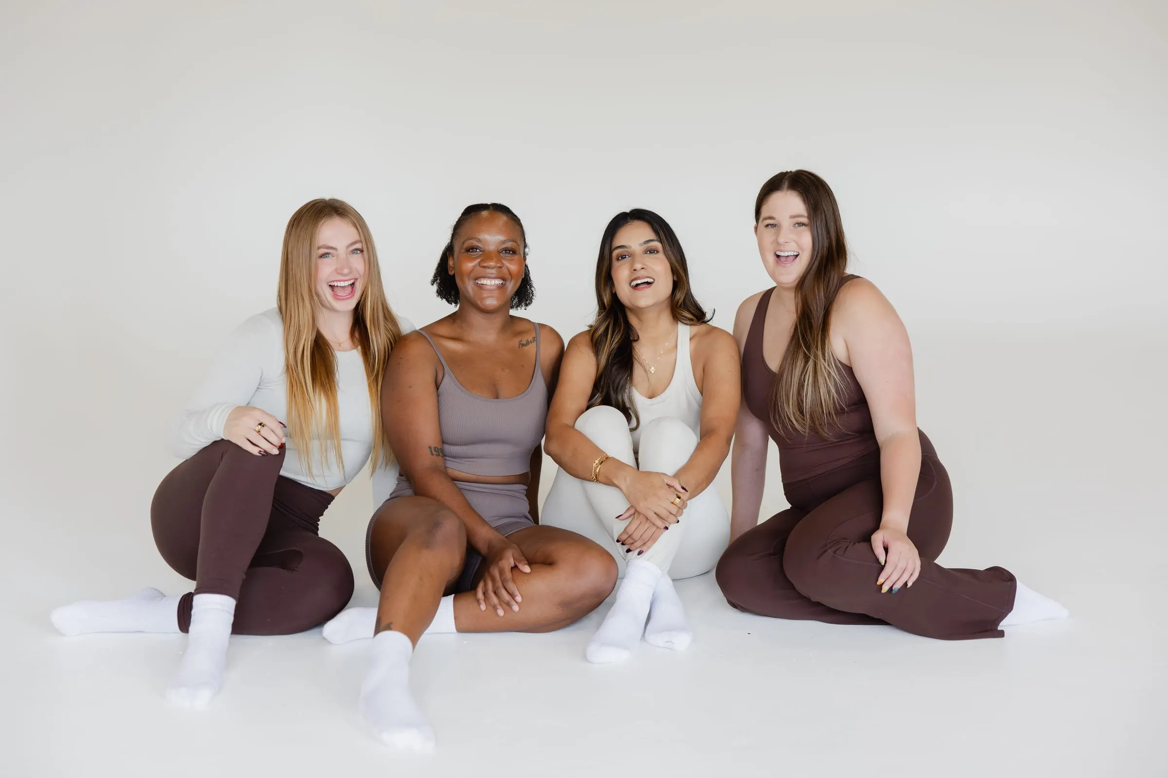 Four women in Pilates wear sitting together on the studio floor, laughing